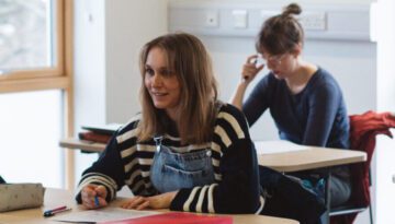 Two students sitting in a classroom; female student in black and white striped shirt is looking up and forwards.