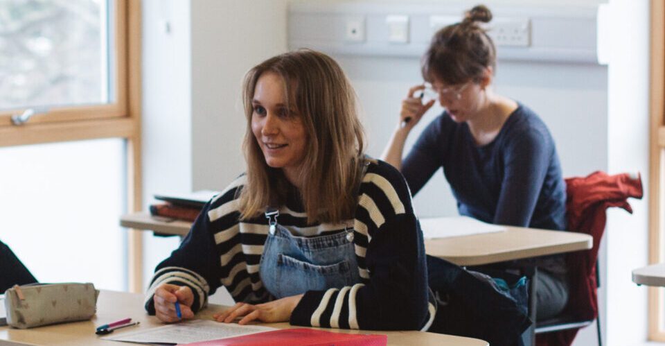 Two students sitting in a classroom; female student in black and white striped shirt is looking up and forwards.